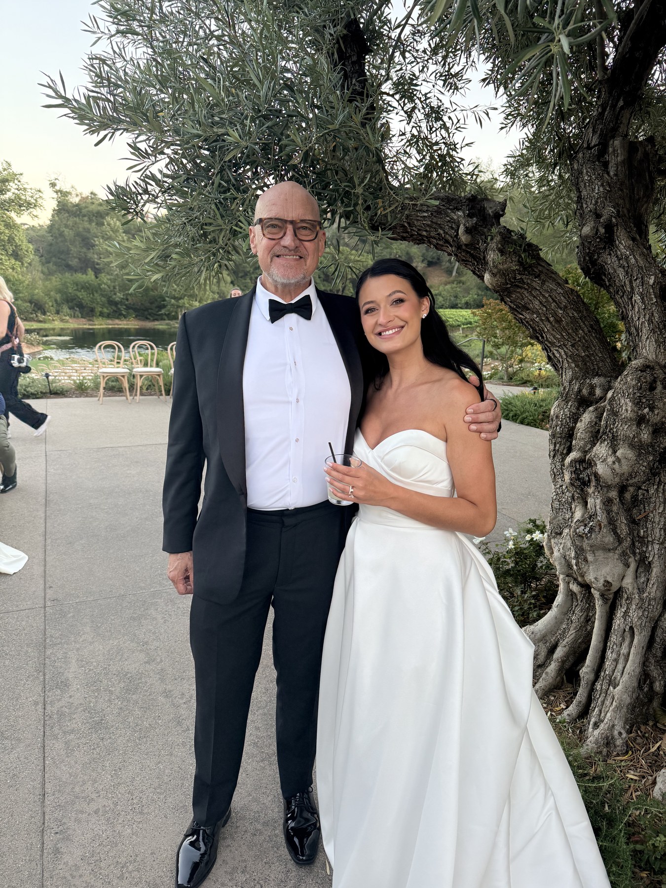Michael in black tie at a wedding, beside the bride, beneath an olive tree.